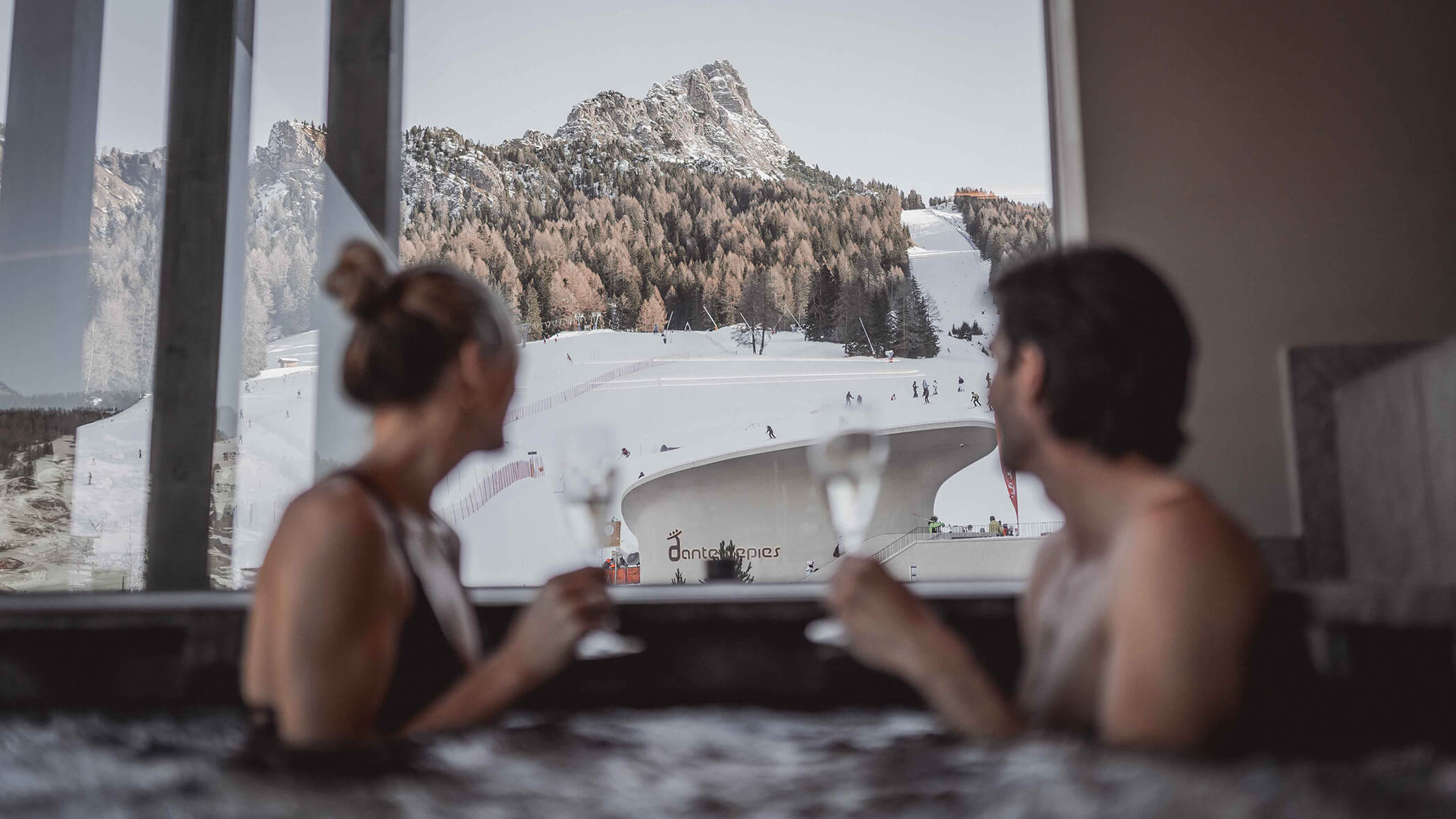Ein Paar blickt mit Prosecco-Gläsern im Pool auf die Skipiste der Sella Ronda - Hotel Continental