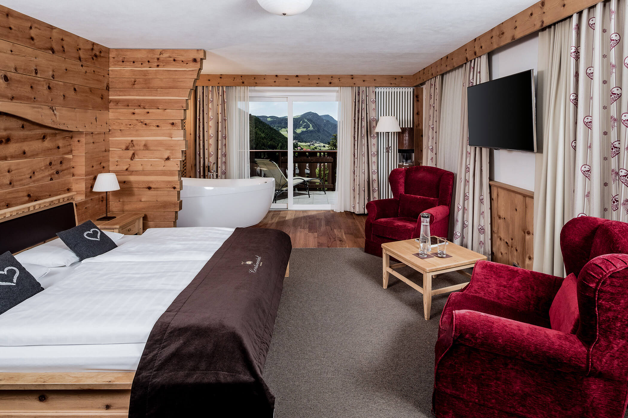 Two red armchairs with a small wooden table on the right and a double bed on the left, with a free-standing bathtub in the background in the Classic Junior Suite 
